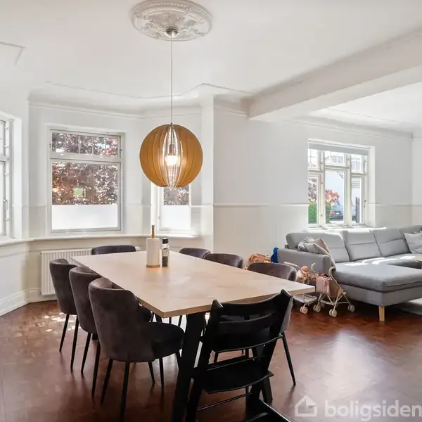 Spisebord med stole er placeret i en lys stue. En stor sofa står i hjørnet ved vinduerne, der giver udsigt til træer udenfor. En lampe hænger over bordet.