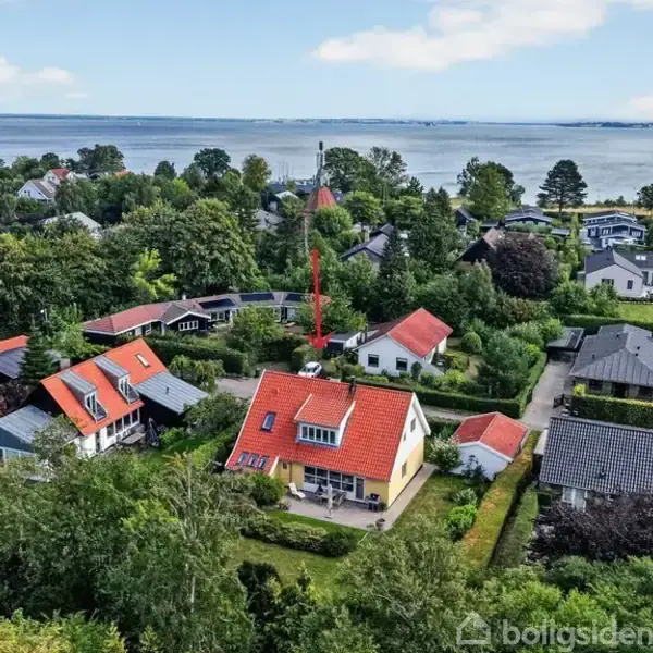 En hvid villa med rødt tag står i en frodig have omgivet af andre huse. Baggrunden viser en kystlinje med hav og skovklædt område.