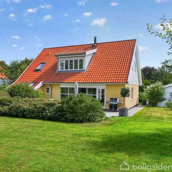 Et gult hus med rødt tag står i en velholdt have med græsplæne og buske. Der er en terrasse med en grill, omgivet af grønne træer og en klar blå himmel.