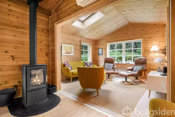 Brændeovn opvarmer hyggelig stue med trævægge; gule og brune møbler står på tæppe ved store vinduer. Billeder hænger på væggen og stor vindue rammer grøn udsigt udenfor.