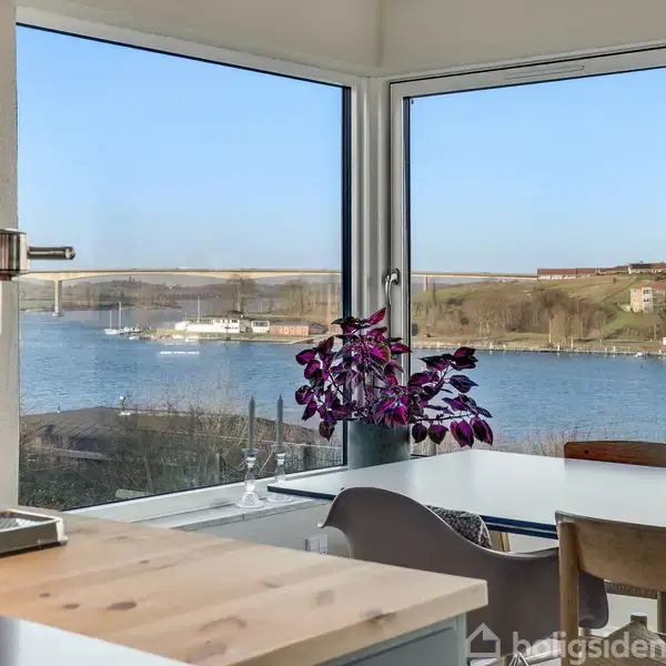 Øen vask i køkkenet har udsigt over en flod med både og en bro i baggrunden. Bord med stole og en hængende lampe giver en indbydende atmosfære.