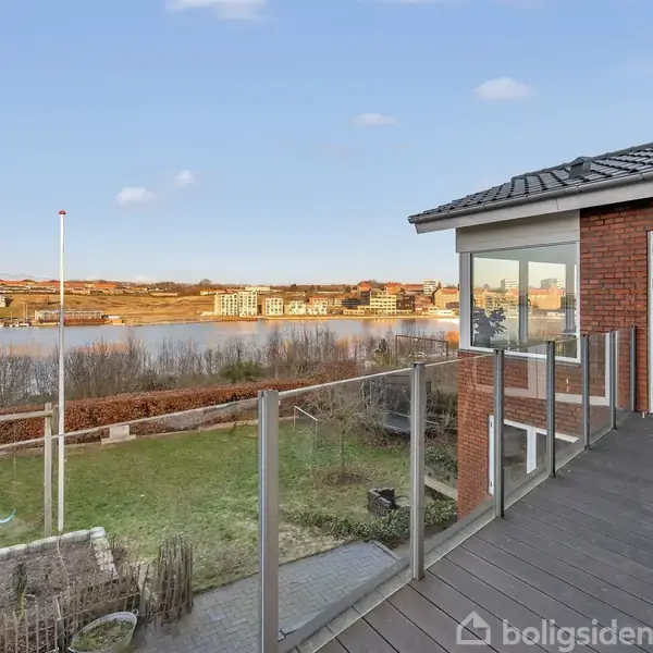 En terrasse med glasrækværk strækker sig fra et murstenshus, der har store vinduer. Udsigt over en sø omgivet af bebyggelser og grønne områder i baggrunden.