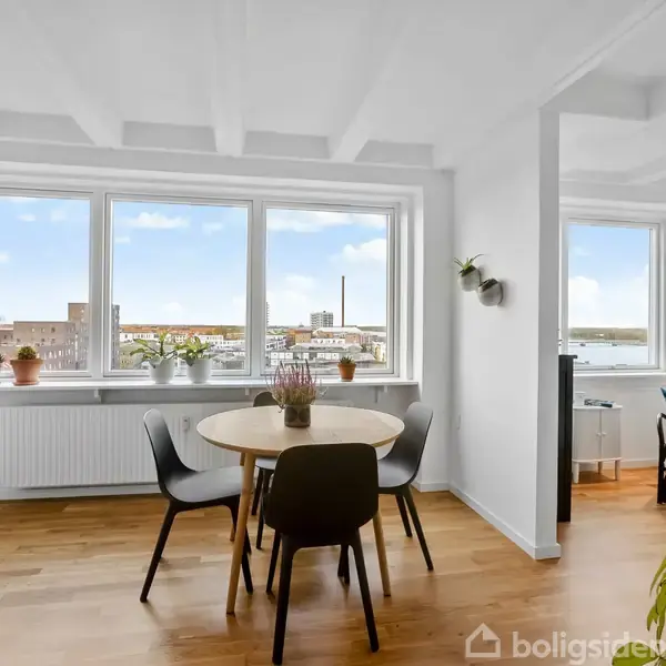 Rundt bord med fire stole står i et lyst rum med store vinduer. Udsigt til byen udenfor, omgivet af planter på vindueskarmen og trælofter.