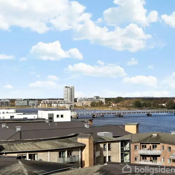 Bygninger står stille med en flod og en bro i baggrunden; omgivet af en klar, blå himmel med spredte skyer.