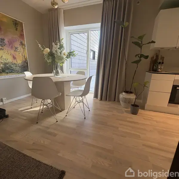 Et rundt bord med en vase blomster står i et lyst rum med trægulv. Fire hvide stole omgiver bordet. Gardiner hænger ved en glasdør, og en plante står ved siden af.