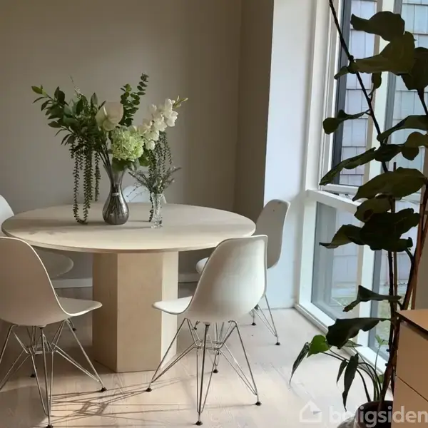 Rundt træbord med blomsterdekoration i glasvase omgivet af hvide stole; placeret ved siden af store vinduer, hvilket giver lys til et moderne, minimalistisk rum.