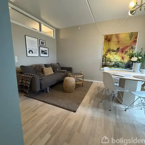 Stue med grå sofa fyldt med puder ved siden af et lille rundt bord og kurv på et tæppe. Væggen har kunstværker over sofaen, og spisebord står ved siden af.