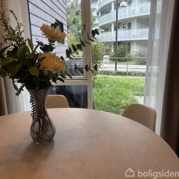 En vase med blomster står på et rundt bord i et lyst rum. Udenfor vinduet ses en græsplæne og et hegn.