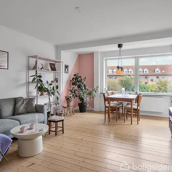 Stue med grå sofa og runde sofaborde, ved siden af en åben reol med planter. Spisebord med træstole ved vinduet, der viser et boligområde udenfor.