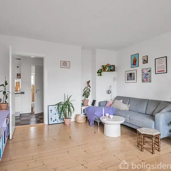 Stue med grå sofa og lille rundt bord, omgivet af indrammede billeder og mange planter. Lyse trægulve og en reol med dekorationer skaber en hyggelig atmosfære.