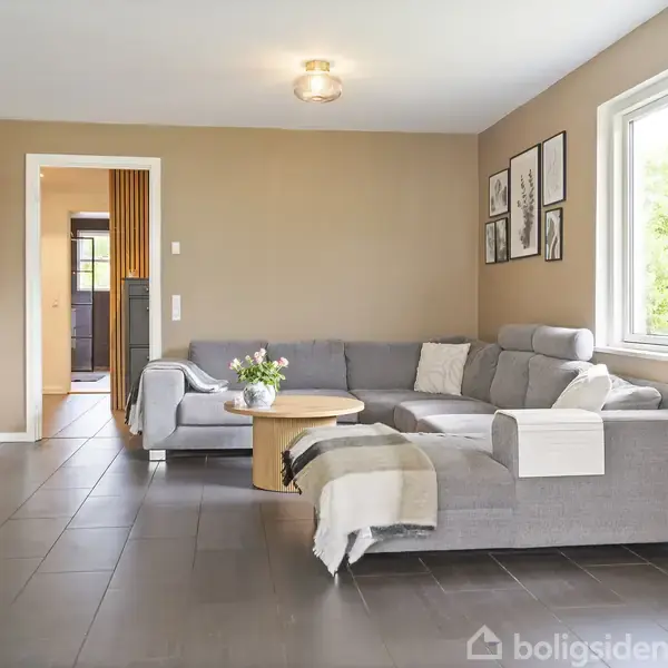 Grå sofa står i stuen ved siden af et træbord med blomster, under et stort vindue med udsigt til en have. Væggene har kunstværker, og en fladskærm hænger til venstre.