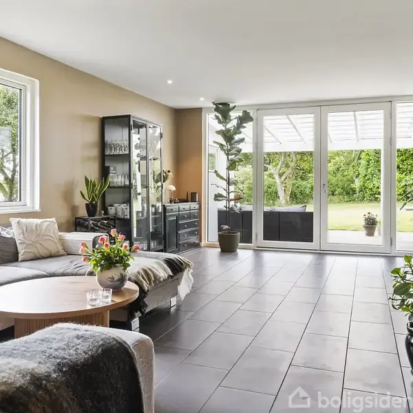 Lædersofa med tæpper og puder står i en lys stue ved et sofabord med blomster, store vinduer åbne til en have med grønne træer og buske.