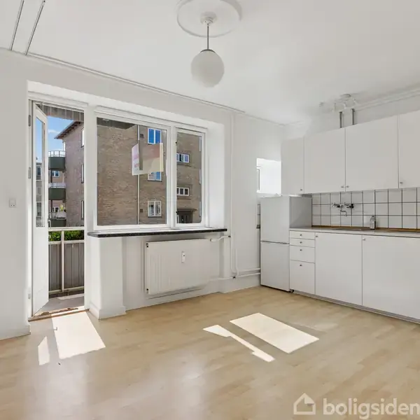 Et lyst køkken med hvide skabe og trælaminatgulv, åbner ud til en lille balkon. Rummet er rigeligt oplyst af dagslys fra et stort vindue og en dør.