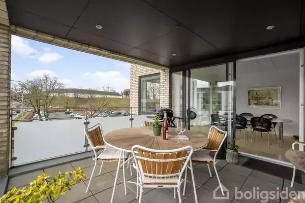 Rundt havebord med flere stole står på altan. Glaspaneler omgiver altanen. Åbne glasdøre forbinder altan med stue, hvor maleri hænger på væggen. Udsigt til parkeringsplads udenfor.