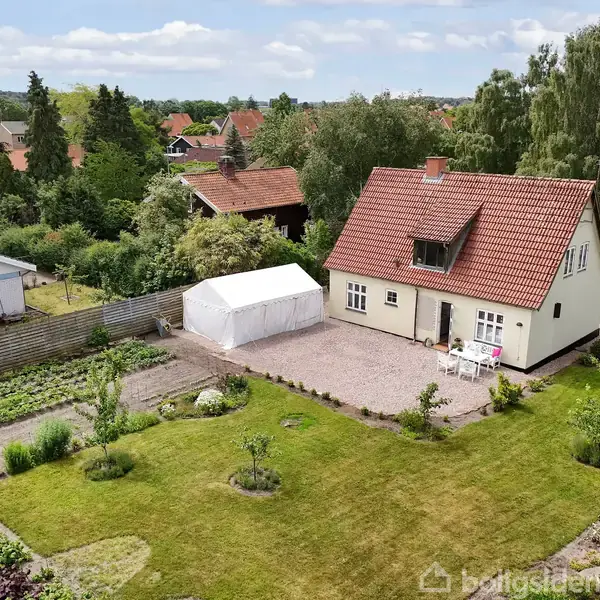 Et rødt taghus med have omgivet af grønne områder og andre huse. Der er en hvid pavillon ved siden af huset, og græsplænen er velplejet med buske og træer.