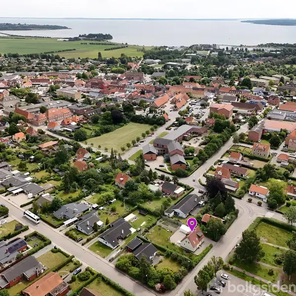 Huse organiserer sig omkring grønne områder, mens veje krydser kvarteret. I baggrunden strækker en sø sig ud mod horisonten, der omfavner landskabet med markante kystlinjer.