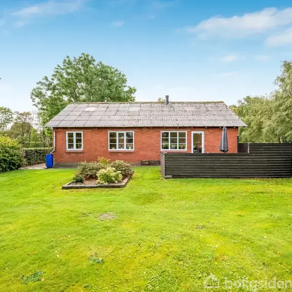 Et rødt murstenshus står stille i en frodig have med græsplæne. Foran huset er en terrasse med parasol og en flagstang. Træer omgiver huset i baggrunden.