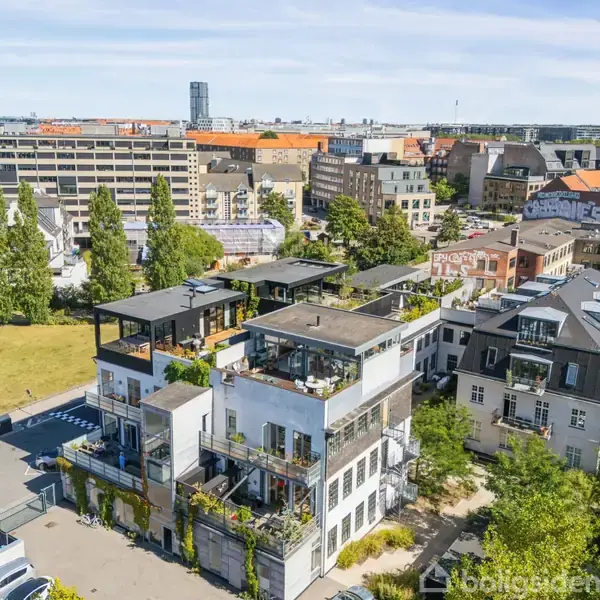 En moderne bygning med flere etager og grønne altaner står i et byområde. Omgivet af træer, nabobygninger og et stort, åbent græsareal. Biler ses på parkeringspladsen.