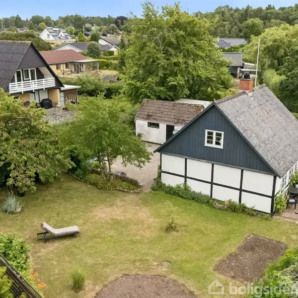 En traditionel bindingsværkshus med stråtag står i en frodig have. Ved siden af er en garage og en liggestol på græsplænen. Omgivet af grønne træer og buske.