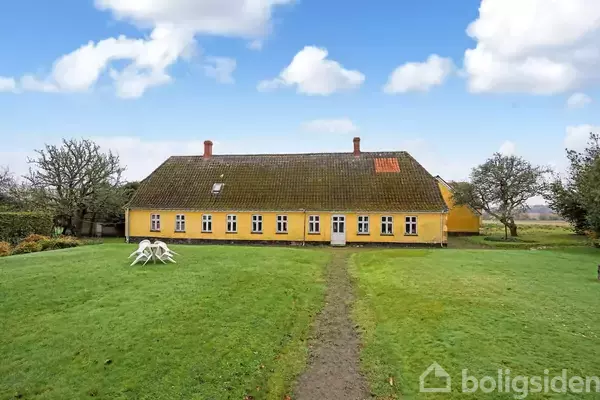 Et gult landsted med skråtag står på en velplejet græsplæne. Der er hvide vinduer, en dør i midten, og et par træstole foran huset.