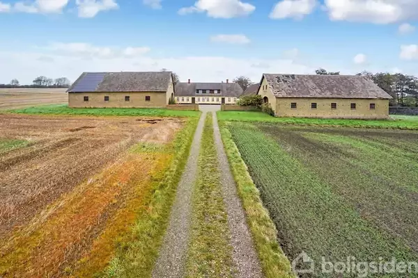To staldbygninger står på hver side af en lang grusvej, der fører mod et hovedhus. Omgivet af marker under en blå himmel med få skyer.