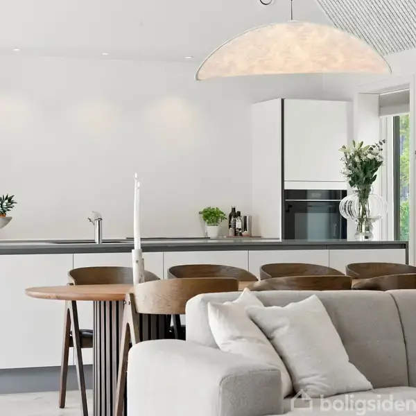 En moderne køkkenindretning med en stor lys lampe hængende over en ø. Barstole står ved øen, og en sofa med hvide puder står foran. Planter og dekorationer er i baggrunden.