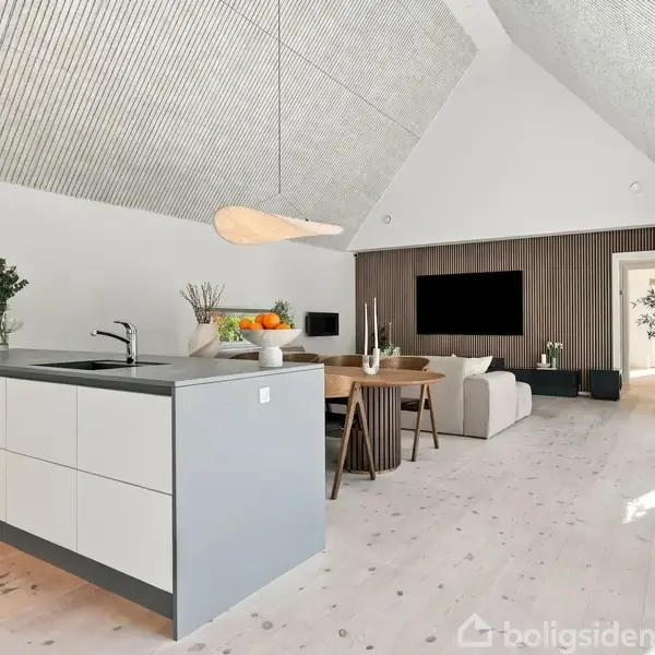 Køkkenø med håndvask og frugtskål står i moderne, lyst alrum, der inkluderer spisebord og stue med fjernsyn. Store vinduer bader rummet i naturligt lys.