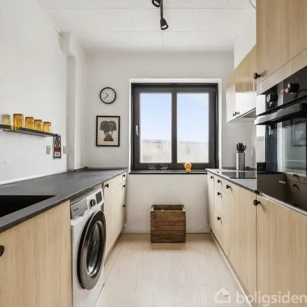 Køkken med træskabe, sort bordplade og vaskemaskine indbygget. I baggrunden ses et vindue, en væguret, hylde med glas og en plakat. Golvet er lyst, rummet er lyst.