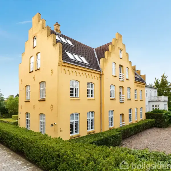 En gul bygning med dekorative gavle står omgivet af velplejede buske og en flisebelagt sti i et grønt område under en klar blå himmel.