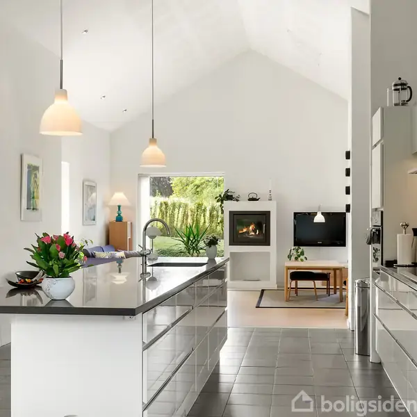 Køkken med glat, hvid køkkenø og forsænkede lamper. Åben stue i baggrunden med sofahjørne og pejs. Lyse fliser på gulvet og grøn udsigt gennem stort vindue.