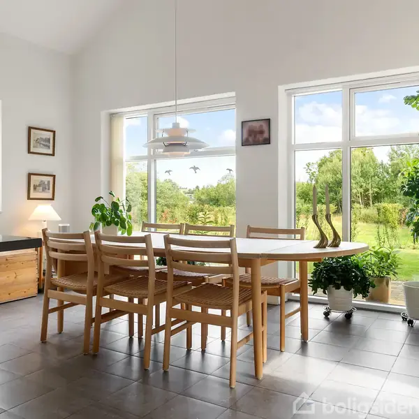 Et spisebord med seks stole står i en lys spisestue. Store vinduer giver udsigt til en grøn have, mens planter og lamper dekorerer rummet.
