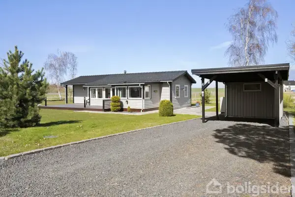 Et enkelt gråt sommerhus med sort tag står stille. Grusvej og lille carport i forgrunden, grøn græsplæne og enkelte træer omkring.