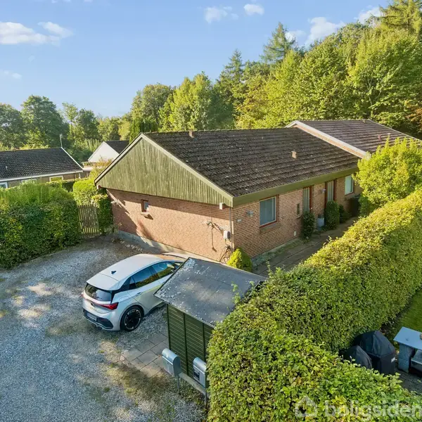 En rød murstensbygning omgivet af grønne hække, med en parkeret hvid bil foran. Grusstier fører gennem en skovfyldt baggrund med flere huse synlige i nærheden.