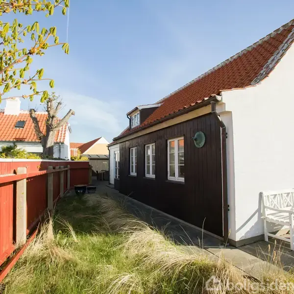 Hus med rødtag og hvide vinduer står ved gangsti. Ved siden af huset er en rød træhegn og en hvid bænk. Omgivet af græs og træer under en solrig himmel.