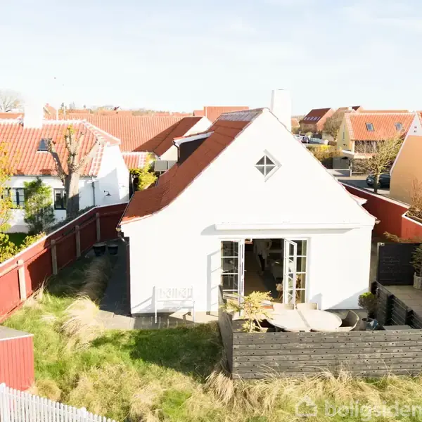 Hvidt hus med rødt tag står omgivet af en have med græs og beplantning. Træhegn indrammer området. Haven har en terrasse med et bord og stole.