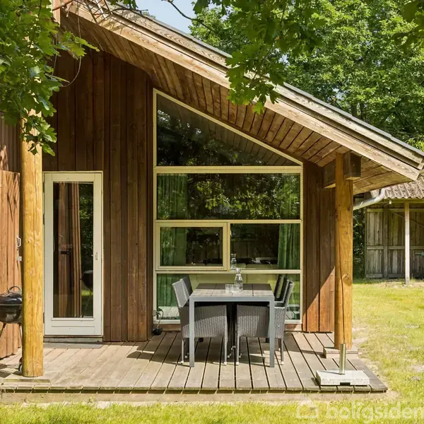 Træhus står stille på en græsplæne med store vinduer og terrasse. Bord med stole ses foran huset, omgivet af træer. I baggrunden er en carport eller udhus.
