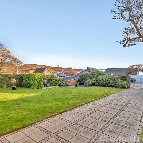 En brolagt sti fører gennem en velplejet have med grønne buske og træer i en forstad. En port afslutter stien med udsigt til en bakke og huse i baggrunden.