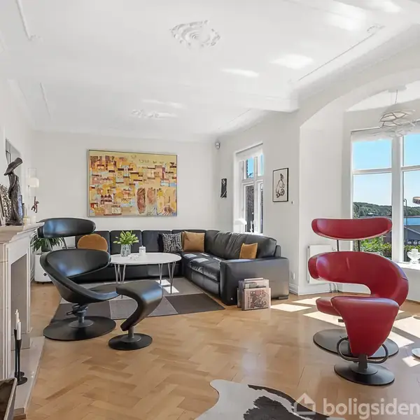 Stue med moderne designstole i sort og rød, lædersofa, kunst på væggene. Store vinduer slipper sollys ind. Parketgulv med tæppe i midten. Elegant og lyst interiør.