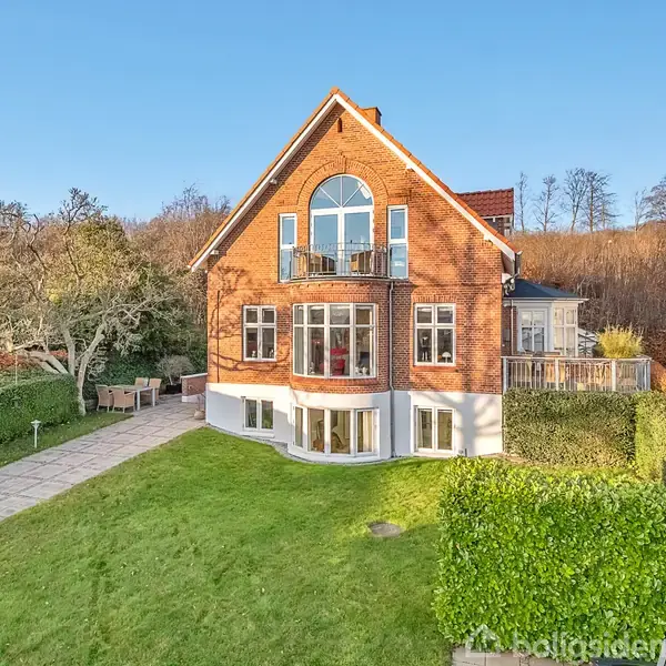 Rødt murstenshus med store vinduer står stille i en solrig have omgivet af velplejede hække og gangstier. En terrasse ses til venstre under et gammelt træ.