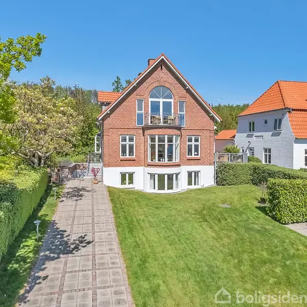 En stor murstensvilla med en rød tegltag står omgivet af en velplejet græsplæne og en fliseterrasse. Ved siden af ligger et lignende hvidt hus.