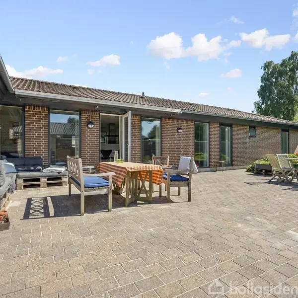 En gårdhave med flisebelægning og flere siddegrupper. Huset i baggrunden er bygget af rød mursten, og der er store vinduer, der åbner ud til terrassen.