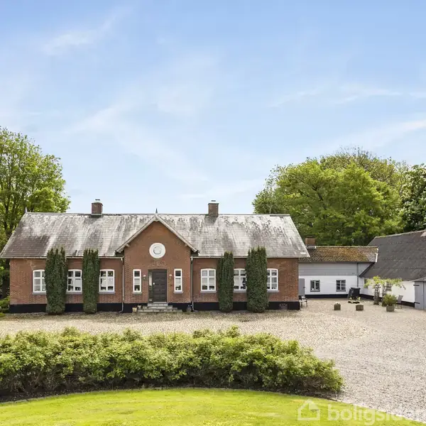 Murstenshus med fem vinduer på hver side af hoveddøren, omgivet af grusgård og grønne buske, flankerede af to fløjesbygninger med stråtag og træer i baggrunden.