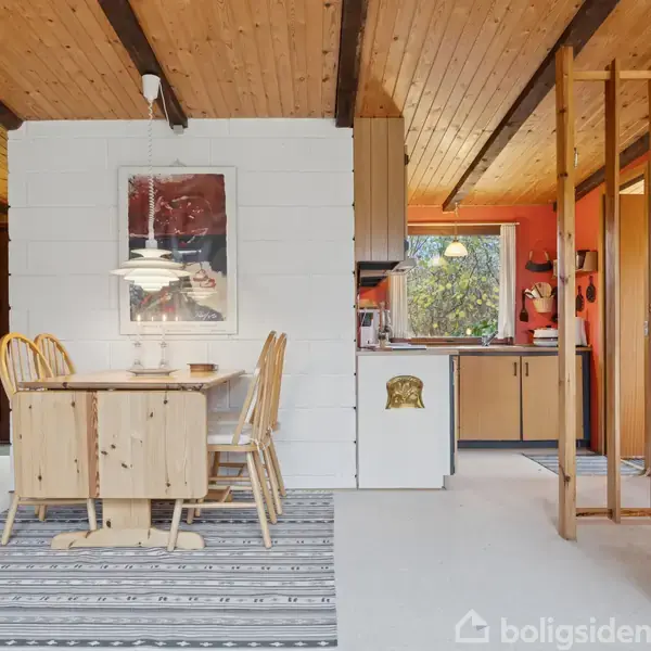 Et lyst træspisebord med stole står i en stue med hvidmalede vægge. Et enkelt køkken ses i baggrunden med et vindue, træpaneler og rødmalede vægge.