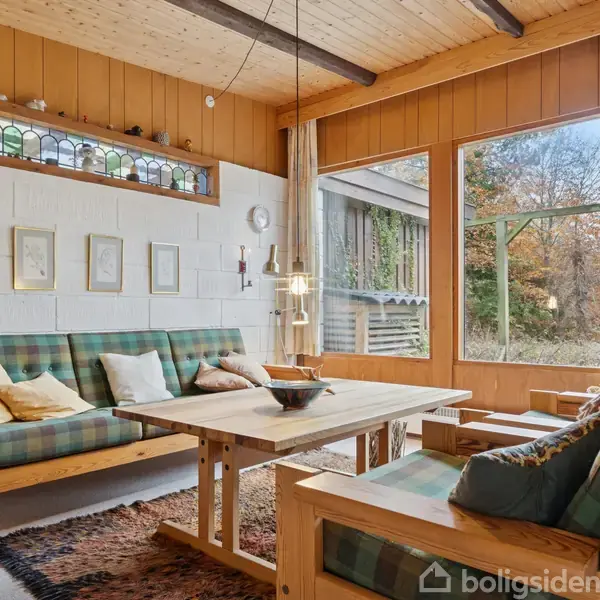 Sofa og lænestol med grønne tern, placeret i en stue med træloft og store vinduer, belyst af en hængelampe. Billeder på væggen og tæppe på gulvet.