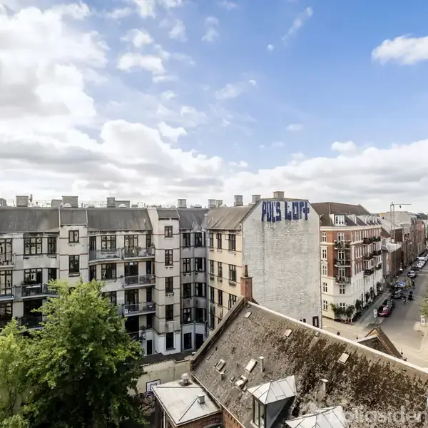En åben vindue udsigt viser en tæt bebygget by med fleretagers lejlighedskomplekser. Der står "ALIS LUF" på en bygning. Skyet himmel ses over hustagene.