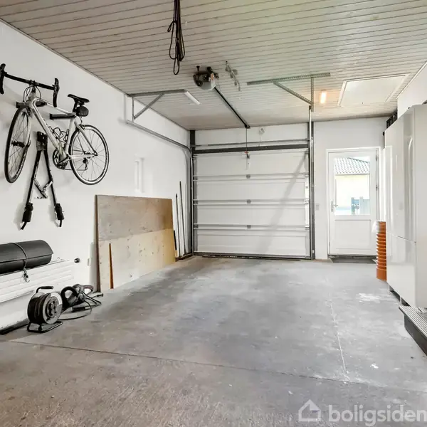 Cykel hænger på væggen i en tom garage med åben port. Ligeledes ses en stige, opbevaringshylder og en fryser i rummet.