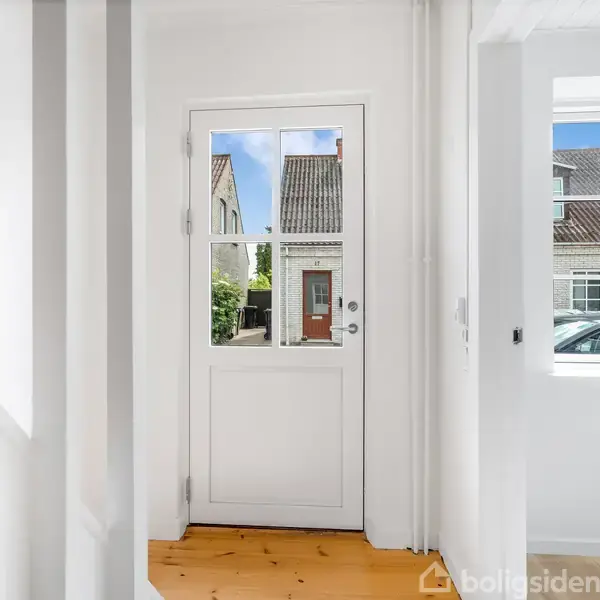 Hvid dør med glasruder står i centrum og leder til en udendørs scene med huse. Lys træplanke gulv omkranser døren. Vindue på højre side viser et bilspejl.