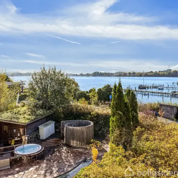 Boblebad og sauna i træ omgivet af frodig natur, placeret nær en søbred med udsigt over både, pontoner og en skov i det fjerne under en klar blå himmel.