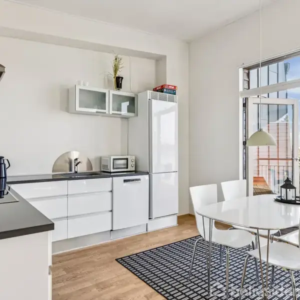 Et moderne køkken med hvide skabe og apparater, hvor en kaffemaskine og mikrobølgeovn står på bordet. Et spisebord med fire stole står ved siden af store vinduer med udsigt til vand og terrasse.