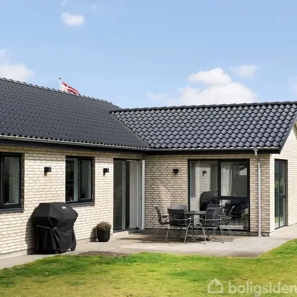 Et moderne hus med lyst murværk står på en græsplæne. En terrasse er udstyret med et bord og stole. Himlen er blå med få skyer.